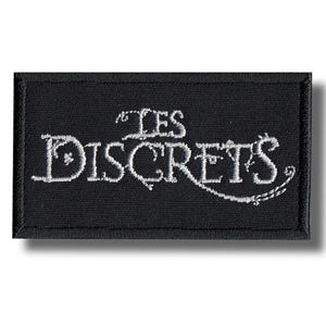 Puede incluir: Parche rectangular negro con texto bordado blanco « LES DISCRETS ». El texto está en una fuente decorativa. El parche tiene un borde negro y una sombra.