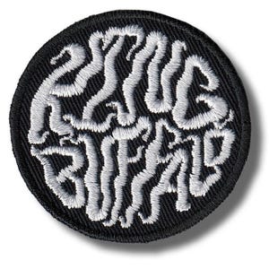 以下が含まれることがあります： 白のスタイリッシュな文字で「ESSAYS BRAIN」と書かれた、丸い黒色の刺繍パッチ。パッチは黒い縁取りがあり、衣類やアクセサリーに縫い付けるようにデザインされています。