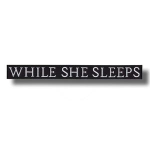 Könnte beinhalten: Ein rechteckiger, schwarzer Aufnäher mit dem weißen, gestickten Text "WHILE SHE SLEEPS". Der Aufnäher ist für das Aufnähen oder Aufbügeln auf Kleidung oder Accessoires gedacht.