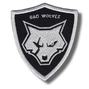 May include: Embroidered patch in a shield shape, featuring a wolf's head design in white against a black background. The text "BAD WOLVES" is arched above the wolf. The patch has a white border.