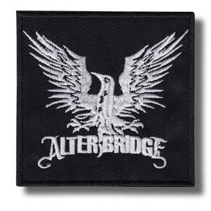 Può includere: Toppa quadrata nera con un'aquila ricamata bianca e il nome della band "ALTER BRIDGE" in un carattere stilizzato. La toppa è probabilmente per una giacca o una borsa.