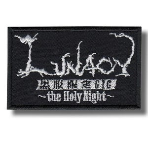 以下が含まれることがあります： 白の刺繍テキストが入った黒い長方形のパッチ。上段には、様式化されたフォントで「Lunacy」と書かれています。その下に別のフォントのテキストがあり、「the Holy Night」というフレーズが一番下にあります。