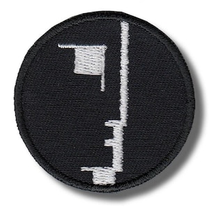 May include: A circular black patch with a white embroidered design. The design features a vertical line with horizontal lines and a small square at the top. The patch has a black border.