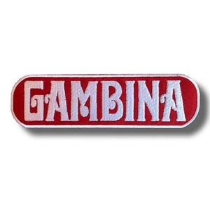 Puede incluir: Parche rectangular rojo con la palabra "GAMBINA" bordada en blanco. Las letras están en una fuente audaz y estilizada. El parche tiene un borde blanco y una ligera sombra.