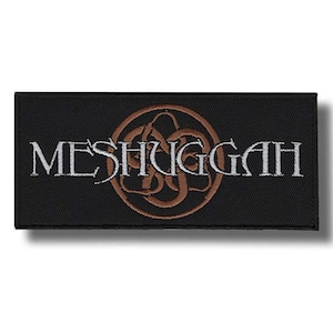 Op de afbeelding: Zwart rechthoekig embleem met het woord "MESHUGGAH" in witte borduursels, met een bruin, gestileerd embleem erboven. Het embleem heeft een getextureerd oppervlak en een lichte schaduw.