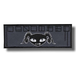 Op de afbeelding: Een rechthoekige, donkergrijze patch met een zwart katachtig wezen met hoorns, witte ogen en hoektanden. Het wezen bevindt zich onder een rij gestileerde, rechthoekige vormen. De patch is waarschijnlijk geborduurd of geweven.
