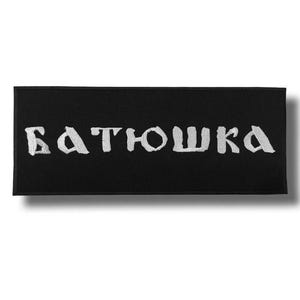 May include: A black rectangular patch with white embroidered text that reads "БАТЮШКА". The patch has a shadow effect, suggesting it is slightly raised. The text is in a stylized, bold font.