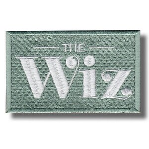 以下が含まれることがあります： セージグリーンの背景に白い文字「THE Wiz」が刺繍された長方形のパッチ。パッチは質感があり、セージグリーンの縁取りが施されています。テキストは様式化されたフォントです。