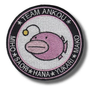 May include: Circular embroidered patch with a pink anglerfish design. The patch has the text "TEAM ANKOU" and names around the fish. The design is on a textured white background, with a black border. Ideal for jackets, bags, or clothing.