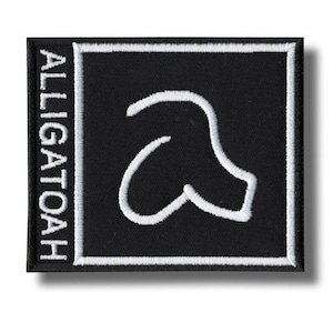 Op de afbeelding: Een vierkante, zwarte geborduurde patch met een witte rand. De patch toont het woord "ALLIGATOAH" verticaal aan de linkerkant en een gestileerde witte "a" in het midden. Het ontwerp is eenvoudig en gedurfd, geschikt voor kleding of accessoires.