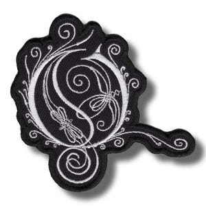 Puede incluir: Parche bordado en blanco y negro con una letra "O" estilizada con diseños florales y remolinos ornamentados. El parche tiene un fondo negro con costuras blancas y una forma única con una extensión curva.