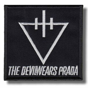 Op de afbeelding: Zwart vierkantige patch met wit geborduurd ontwerp en tekst. Het ontwerp toont een naar beneden wijzende driehoek met een verticale lijn en twee horizontale lijnen erboven. De tekst "THE DEVIL WEARS PRADA" staat eronder.