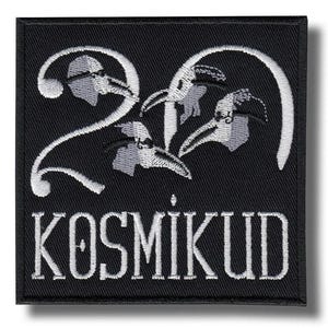 Peut inclure: Écusson carré noir avec texte et motif brodés en blanc. Le motif représente le chiffre "20" avec trois figures stylisées de médecins de la peste. Le mot "KOSMIKUD" est brodé en dessous.