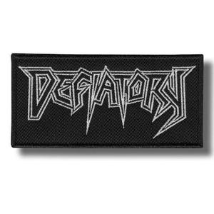 以下が含まれることがあります： 黒い長方形のパッチ。白で「DEFATOKS」の文字が刺繍されており、様式化されたゴシック体の文字です。パッチには黒い縁取りがあり、衣類やバッグ用のアクセサリーである可能性があります。