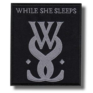 Op de afbeelding: Zwart geborduurde patch met de tekst "WHILE SHE SLEEPS" boven een gestileerd grijs logo. Het logo is een combinatie van de letters "W" en "S". De patch is rechthoekig met een zwarte rand.