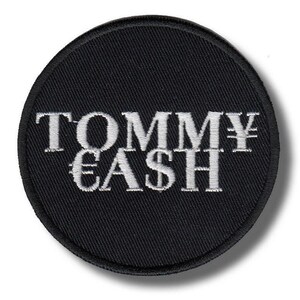 Op de afbeelding: Een ronde, zwarte geborduurde patch met de tekst "TOMMY CASH" in witte letters. Het ontwerp bevat een yen-symbool en een euro-symbool. De patch heeft een zwarte rand.