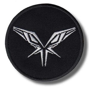 May include: A circular, black embroidered patch with a white, symmetrical design, possibly wings or a geometric butterfly. The patch has a black border and a textured fabric appearance.