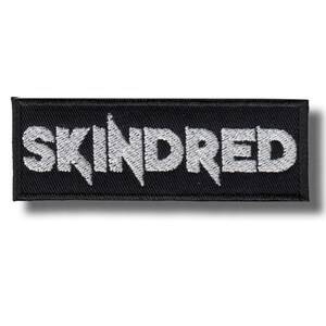 Può includere: Una toppa rettangolare nera con la scritta "SKINDRED" ricamata in bianco. Il carattere è stilizzato. La superficie della toppa è strutturata, probabilmente in tessuto.