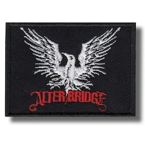 Pode incluir: Patch retangular preto com uma águia branca com as asas abertas. O texto vermelho "ALTER BRIDGE" está bordado abaixo. O patch tem uma borda preta.