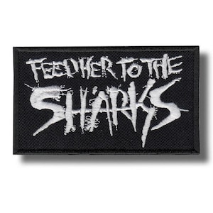 Può includere: Toppa rettangolare nera con la scritta bianca "FEED HER TO THE SHARKS" in un carattere gotico invecchiato. La toppa è progettata per essere cucita su vestiti o borse, con un bordo nero.