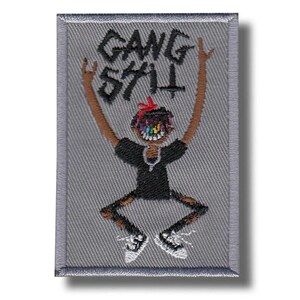 Op de afbeelding: Geborduurde patch met een grijze achtergrond en een cartoonfiguur met opgeheven armen. De figuur heeft een bruine huid, een zwart shirt en een gescheurde broek. De tekst "GANG 574" staat boven de figuur.