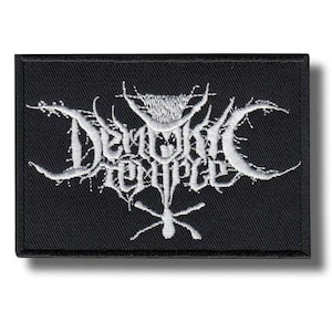 May include: Black rectangular patch with white embroidered text that reads "Demonic Temple" in a gothic font. The patch has a detailed, stylized design with sharp edges and a cross-like symbol at the bottom.