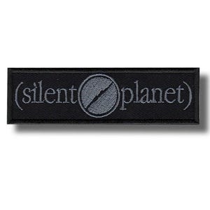 May include: Black rectangular patch with the text "(silent planet)" embroidered in gray thread. A gray circle with a diagonal line through it is centered between the words. The patch has a black border.