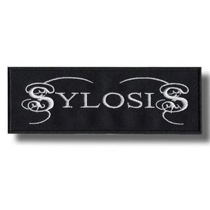 Puede incluir: Parche rectangular negro con la palabra "SYLOSIS" bordada en blanco, con una fuente estilizada. El parche tiene un borde decorativo blanco encima y debajo del texto. El parche es probablemente para ropa o accesorios.