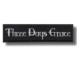 Może przedstawiać: Prostokątna czarna naszywka z wyhaftowanym białym napisem "Three Days Grace" w gotyckim stylu. Naszywka ma czarną obwódkę i prawdopodobnie jest przeznaczona do przyszywania do odzieży lub akcesoriów.