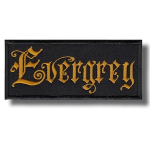 May include: A black rectangular patch featuring the word "Evergrey" in gold gothic script. The patch is embroidered with a black border. The design is simple and bold, suitable for clothing or accessories.