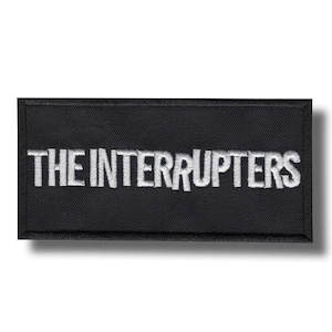 Może przedstawiać: Czarna prostokątna naszywka z białym haftowanym napisem "THE INTERRUPTERS". Naszywka przeznaczona do umieszczenia na odzieży lub akcesoriach, prezentująca nazwę zespołu.