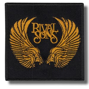 May include: A square black patch featuring the words "RIVAL SONS" in gold lettering, framed by a gold winged design. The patch is likely for clothing or accessories.