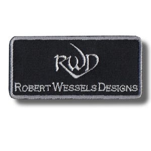 Peut inclure: Un écusson rectangulaire noir brodé avec une bordure grise. L'écusson présente le texte brodé blanc "RWD" dans une police stylisée au-dessus des mots "ROBERT WESSELS DESIGNS" en blanc.