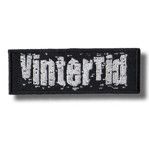 Op de afbeelding: Zwart rechthoekig embleem met het woord "Vintertid" in witte, gotische letters geborduurd. Het embleem heeft een zwarte rand en een getextureerde uitstraling. Het ontwerp is eenvoudig en gedurfd, geschikt voor kleding of accessoires.