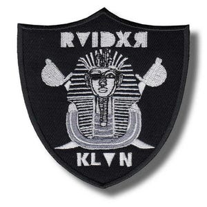 Peut inclure: Écusson noir en forme de bouclier avec texte et motif brodés en blanc. Le motif représente une tête de pharaon avec des outils croisés et le texte "RVIDXR" au-dessus et "KLVN" en dessous. L'écusson est probablement pour les vêtements ou accessoires.