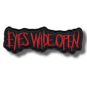 May include: A black embroidered patch with the words "EYES WIDE OPEN" in red, stylized lettering. The patch has a jagged, irregular border. The text appears to be stitched onto the black fabric.