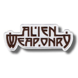 May include: Embroidered patch with the words "ALIEN WEAPONRY" in brown lettering on a white background. The patch has a rectangular shape with rounded edges and a white border. The patch is designed to be ironed on or sewn onto fabric.