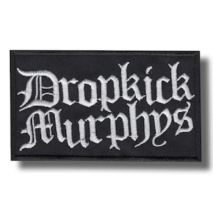 Może przedstawiać: Czarna prostokątna naszywka z wyhaftowanym napisem "Dropkick Murphys" w białych gotyckich literach. Naszywka ma czarną obwódkę i prawdopodobnie jest przeznaczona do przyszywania do odzieży lub akcesoriów.