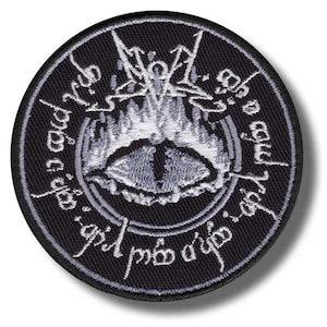 May include: Round black patch with a white embroidered design featuring an eye, flames, and script. The eye is in the center, surrounded by flames and script. The patch has a white border.