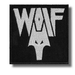 May include: Square black patch with white embroidered text and a wolf head design. The text reads "WAF" with the wolf head incorporated into the design. The patch has a white border and a shadow effect.