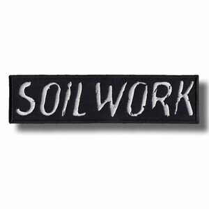 Puede incluir: Parche rectangular negro con la palabra "SOILWORK" bordada en blanco, con letras estilizadas. El parche tiene un borde negro y probablemente está diseñado para ser cosido o planchado en ropa o accesorios.