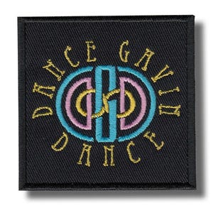 May include: Black square patch with embroidered text and design. The design features a central blue and pink double-circle with a vertical blue line through the center. The text "DANCE GAVIN DANCE" is embroidered in gold around the circle.