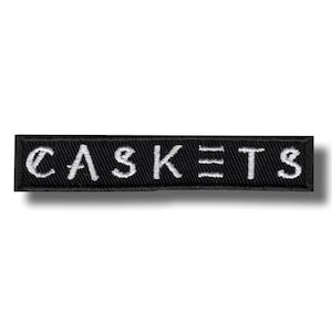 May include: A black rectangular embroidered patch featuring the word "CASKETS" in white. The text is in a simple, sans-serif font, and the patch has a clean, minimalist design. The word is centered.