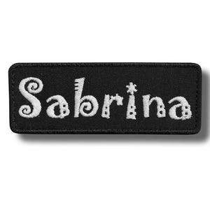 Może przedstawiać: Czarna, prostokątna naszywka z wyhaftowanym imieniem "Sabrina" w kolorze białym. Litery mają fantazyjną, dekoracyjną czcionkę. Idealna do odzieży lub akcesoriów.