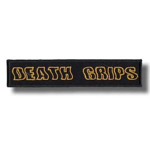 May include: A rectangular black patch with the words "DEATH GRIPS" embroidered in gold-colored thread. The patch has a simple, clean design, with the text centered and the letters outlined.