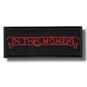 Könnte beinhalten: Ein rechteckiger, schwarzer Aufnäher mit dem gestickten Text "IN THIS MOMENT" in Rot. Der Text ist mit dekorativen Verzierungen versehen. Der Aufnäher ist wahrscheinlich für Kleidung oder Accessoires.