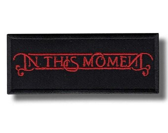 May include: A rectangular black patch with the embroidered text "IN THIS MOMENT" in red. The text is stylized with decorative flourishes. The patch is likely for clothing or accessories.