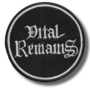 Op de afbeelding: Een ronde, zwarte patch met witte geborduurde tekst "Vital Remains" in een gotisch lettertype. De patch heeft een witte rand en is waarschijnlijk voor kleding of accessoires.