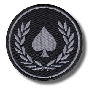 Op de afbeelding: Een ronde, zwarte patch met een grijs schoppensymbool in het midden, omringd door een grijze lauwerkrans. De patch heeft een grijze rand en is waarschijnlijk een geborduurd embleem.