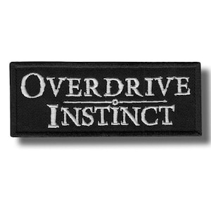 Op de afbeelding: Zwart rechthoekig embleem met witte geborduurde tekst "OVERDRIVE INSTINCT". De patch heeft een zwarte rand en een schaduweffect. De tekst is in een vetgedrukte, schreefloze lettertype.